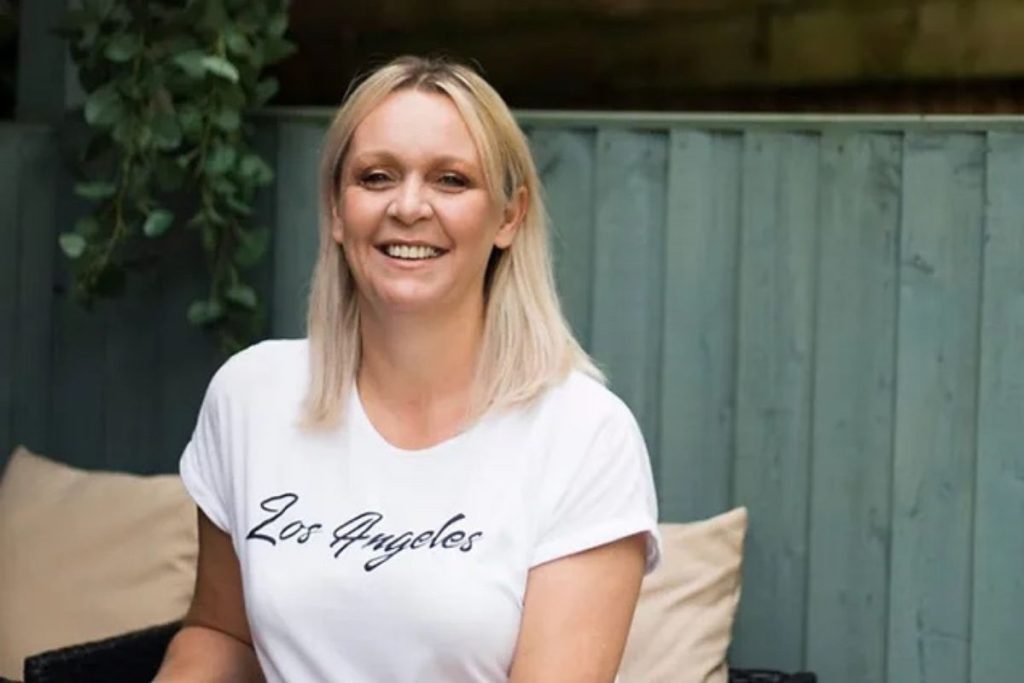 A blonde woman wearing a white t-shirt that says "Los Angeles" smiles brightly while sitting outdoors against a light blue wooden fence and surrounded by cushions and greenery.