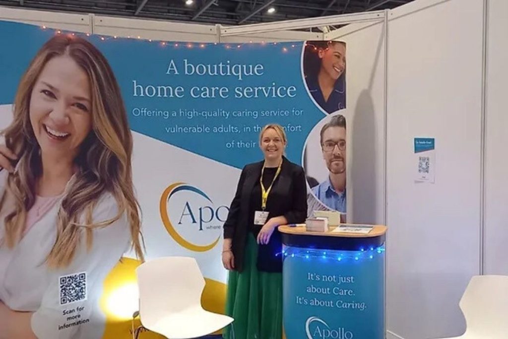 A woman in a black jacket and long green skirt stands smiling at an exhibition booth for Apollo, a boutique home care service, which features a large banner and a small circular table.