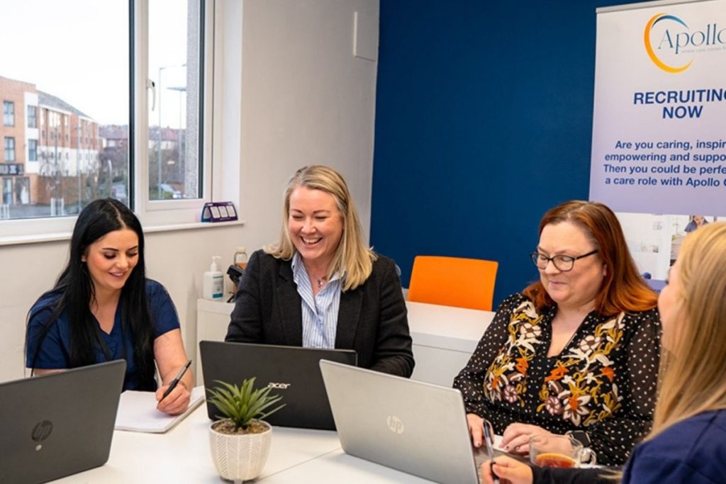Four women are seated around a white conference table with laptops, a notepad, and a small potted plant, working together in a bright office with a blue accent wall and an Apollo recruiting banner.