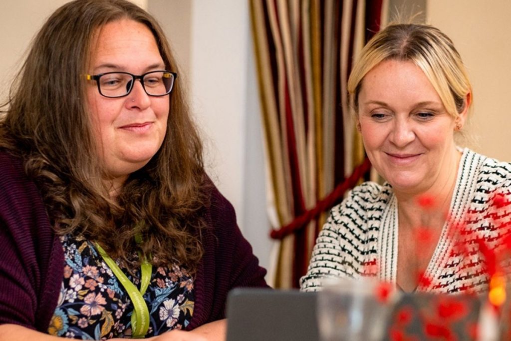Two women smile while looking down at a tablet or screen. One has long dark hair and glasses and wears a floral top; the other has blonde hair and wears a white and black patterned sweater.