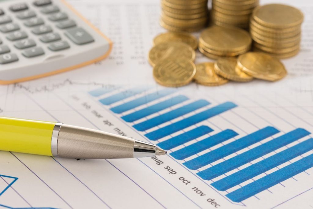A close-up of a paper chart showing blue bar graphs and line graphs, with a silver and yellow pen tip resting on the data. In the blurred background are a calculator and several stacks of gold coins, representing financial analysis and benefits.