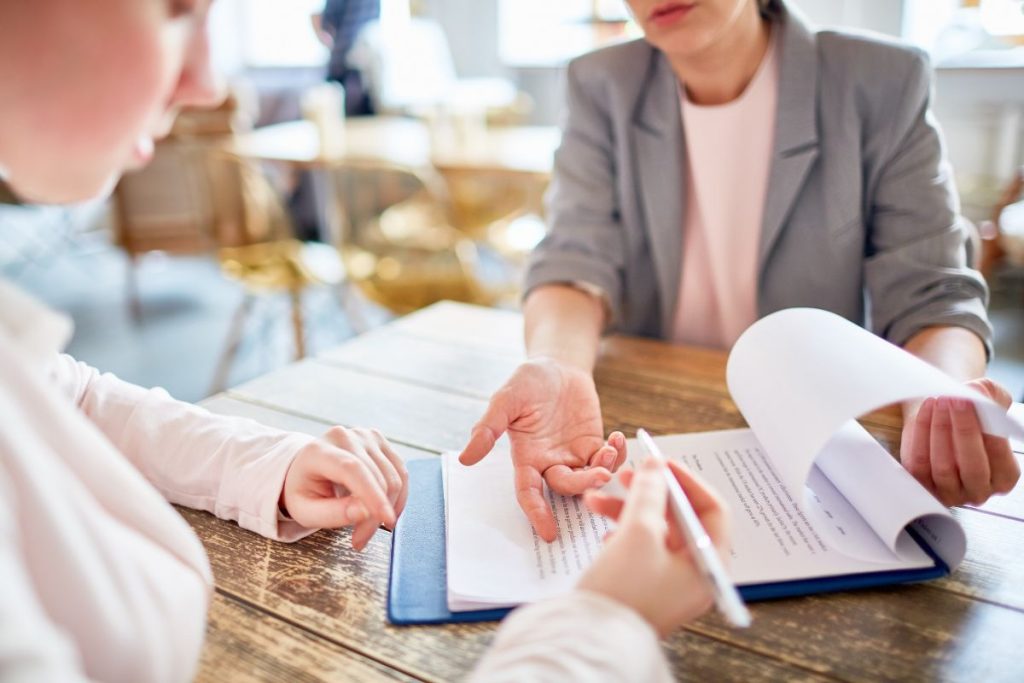A close-up of two people at a wooden table reviewing a contract or business document together. One person in a gray blazer is pointing to text on the page while the other person holds a pen ready to sign, suggesting a business discussion or long-term agreement.