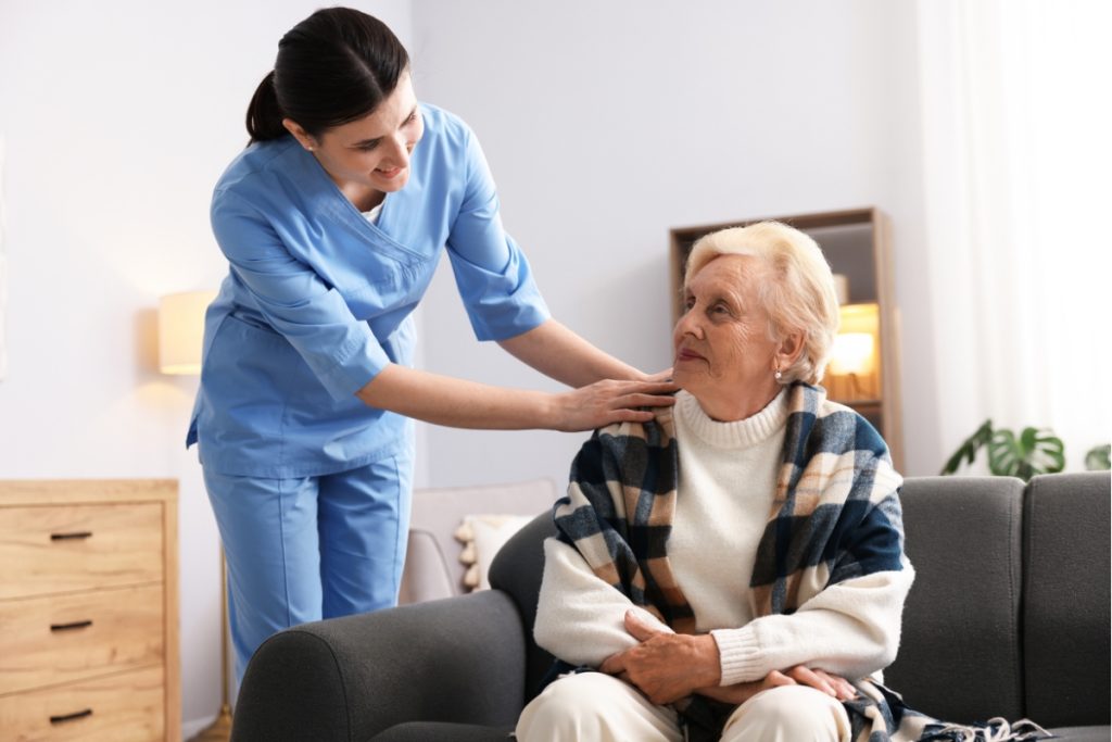 A caregiver in blue scrubs smiling warmly while placing a supportive hand on the shoulder of an elderly woman sitting on a sofa.