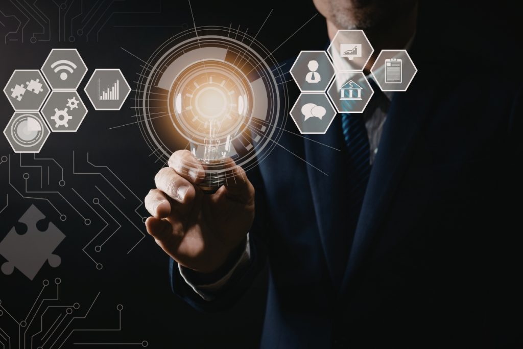 A businessman in a dark suit holding a glowing lightbulb, surrounded by digital icons representing technology, finance and data.