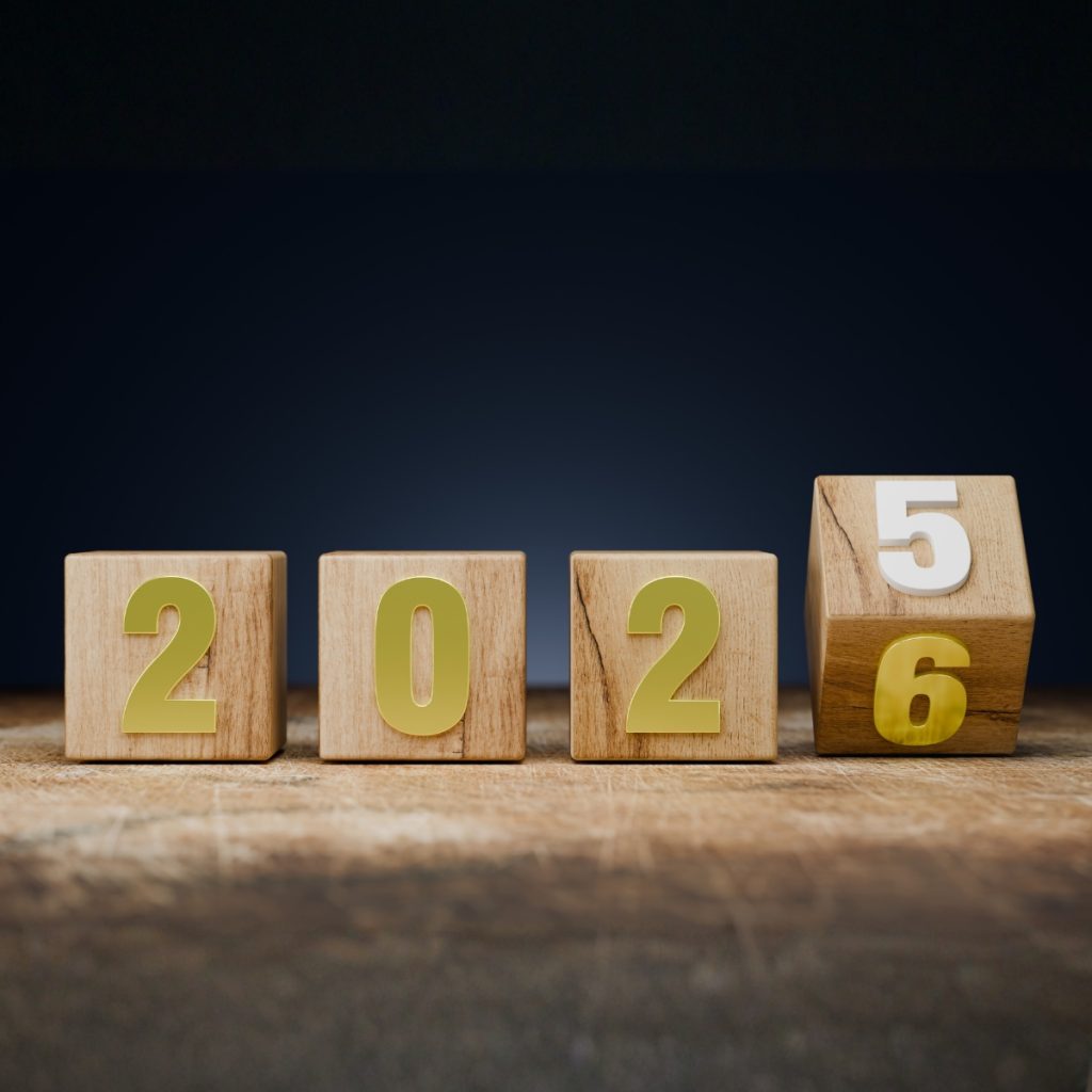 Four wooden blocks on a surface; the first three show "202," and the fourth is rotating to change the year from 2025 to 2026.