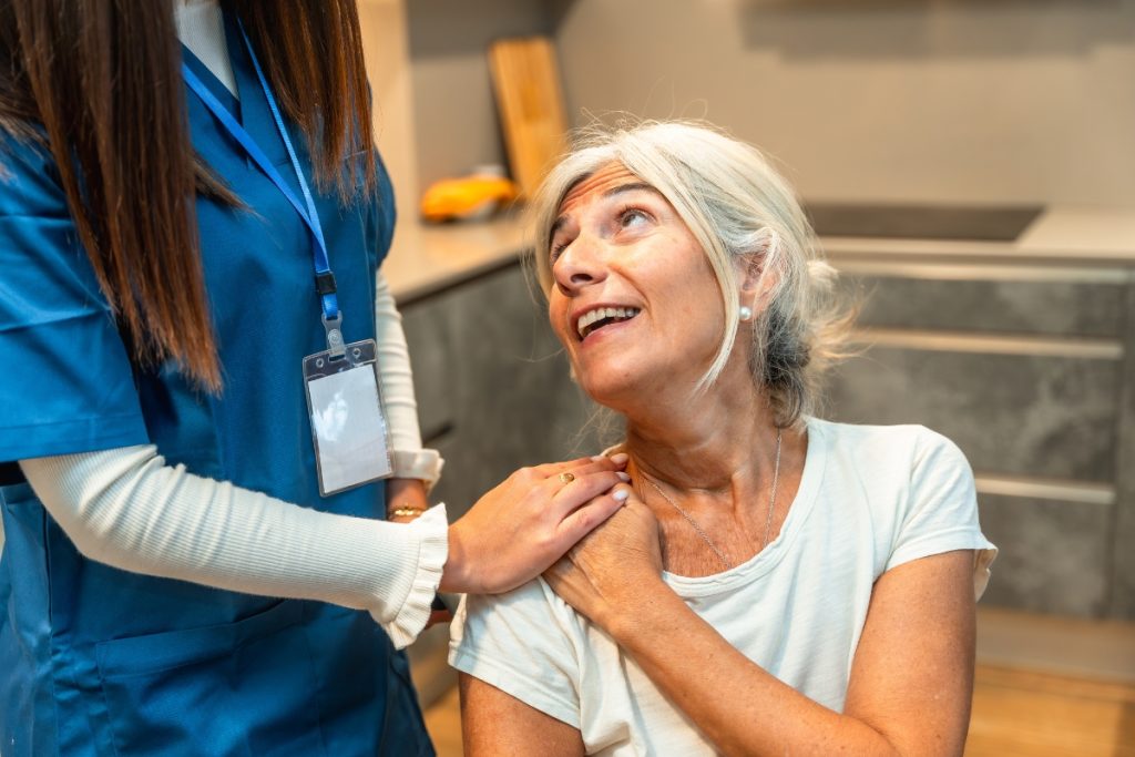 A caregiver in blue scrubs resting a hand on the shoulder of a smiling elderly woman in a kitchen.