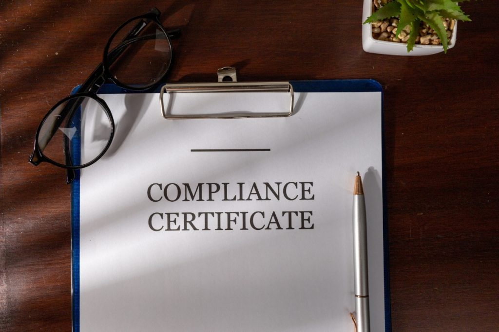 A clipboard on a wooden desk holding a document titled "COMPLIANCE CERTIFICATE" next to a pen and glasses.