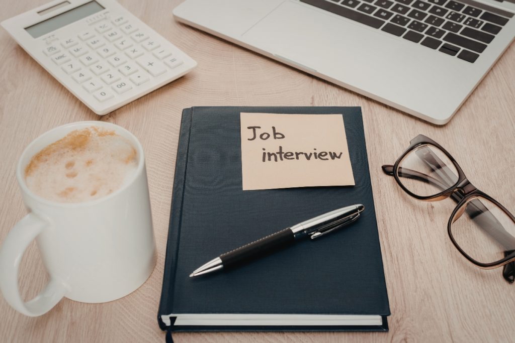 A notebook with a sticky note reading "Job interview" on a desk with a laptop, calculator, and coffee.