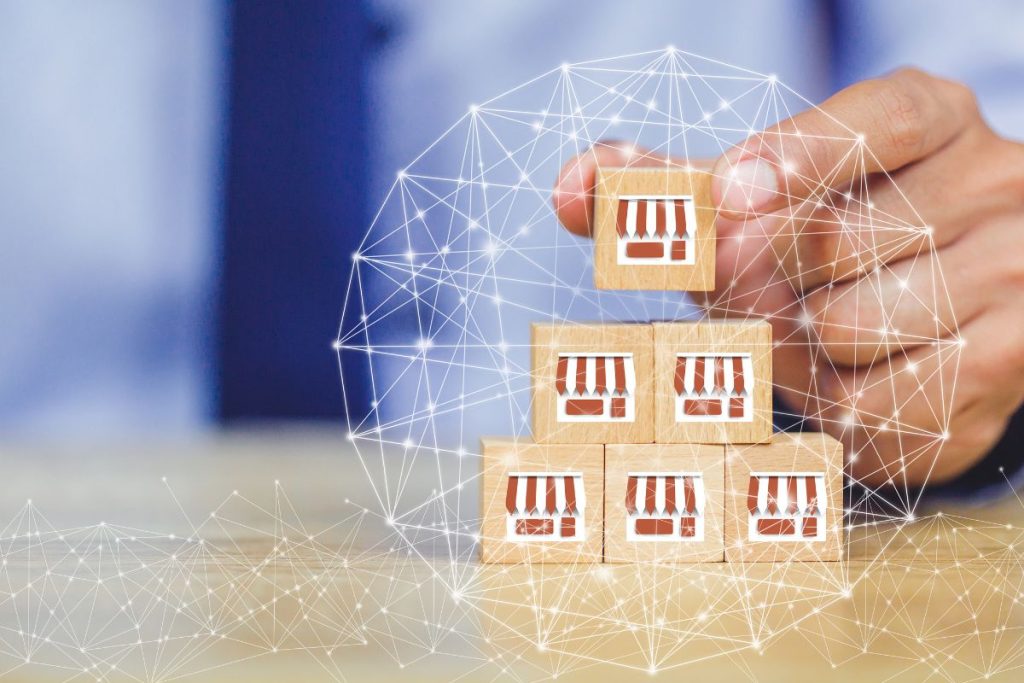 A person’s hand placing a wooden block with a storefront icon on top of a pyramid of similar blocks, overlaid with a glowing white digital network web.